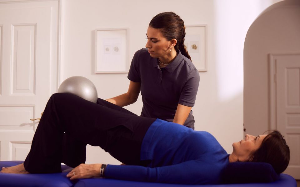 Physiotherapeutin unterstützt Patientin bei Übung mit Gymnastikball im Raum.