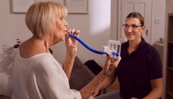 Ältere Frau übt Atemtherapie mit Gerät, betreut von Therapeutin.