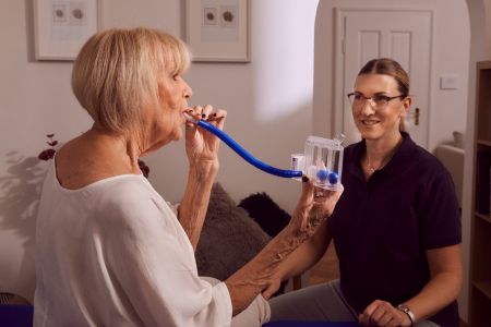 Physiotherapie und Atemtherapie bei Lungenerkrankter Patientin im Hausbesuch, die Patientin trainiert mit einem Atemtrainer zu Hause und wird von der Atemtherapeutin angeleitet.