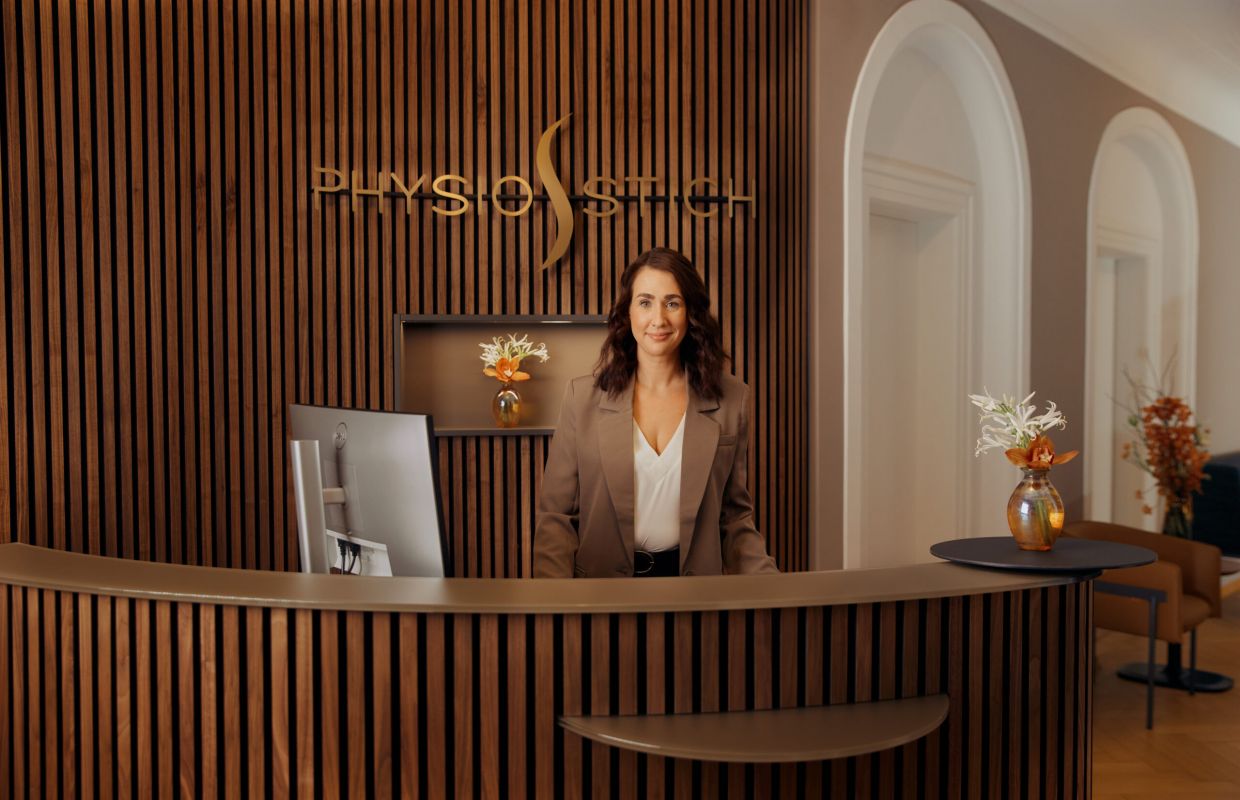 Inhaber und Unternehmer einer privaten Physiotherapiepraxis, welche auf Hausbesuche in Hamburg, Berlin, Frankfurt, Wiesbaden, Mainz, Taunus, München, Bonn, Köln und Düsseldorf spezialisiert ist. Die stationäre Praxis befindet sich in München-Schwabing. Der Chef der Physiotherapiepraxis sitzt auf einer blauen, mobilen Physiotherapiebank bei einem Patienten zu Hause in der Wohnung, trägt eine beige Hose und einen blauen Pullover. Er lächelt in die Kamera.