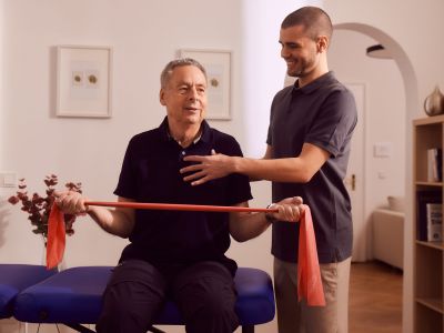 Älterer Mann trainiert mit Theraband, Physiotherapeut assistiert, in Praxis.