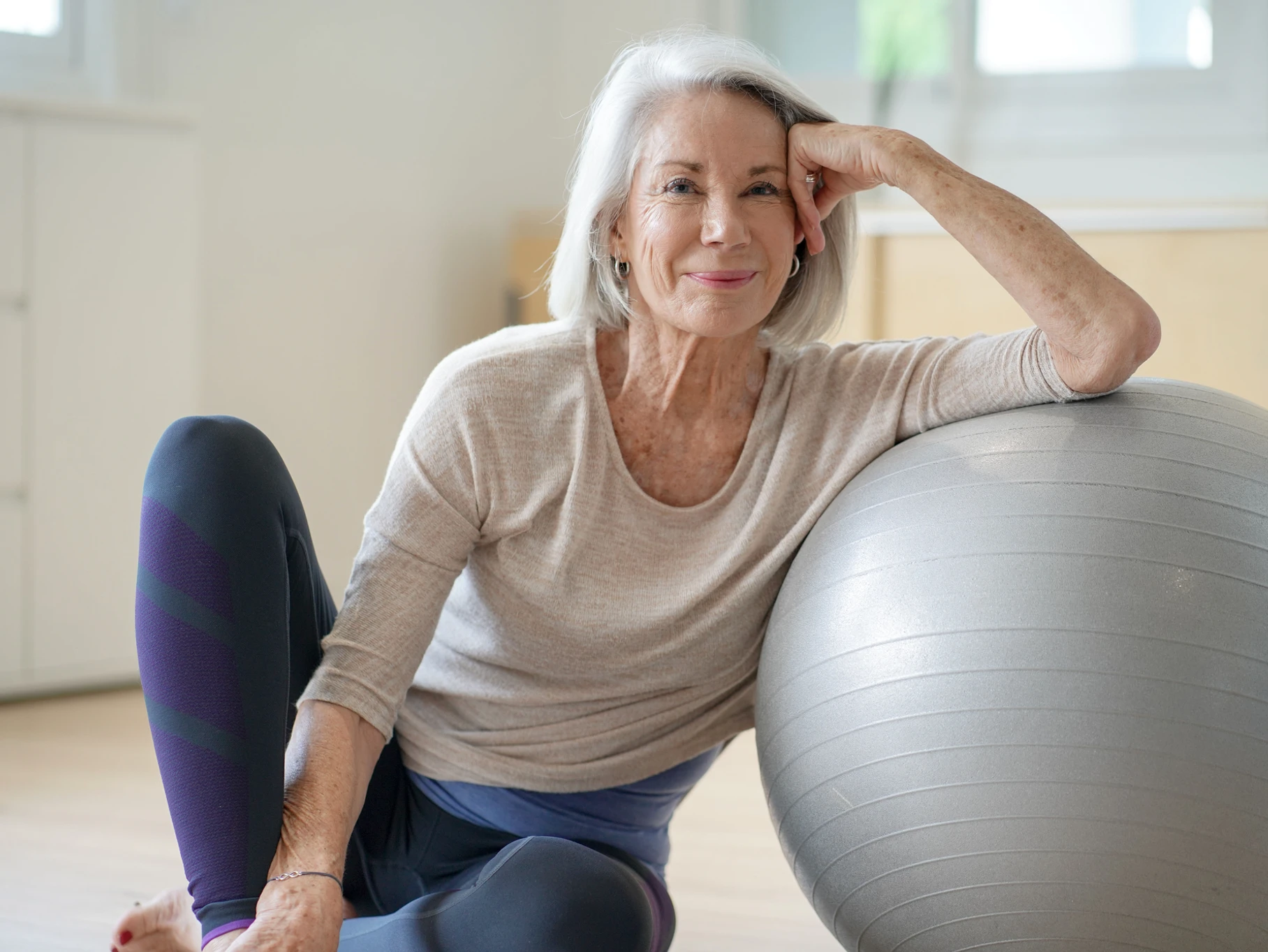 Ältere Frau sitzt auf dem Boden eines Trainingsraums und lehnt sich an einen Gymnastikball