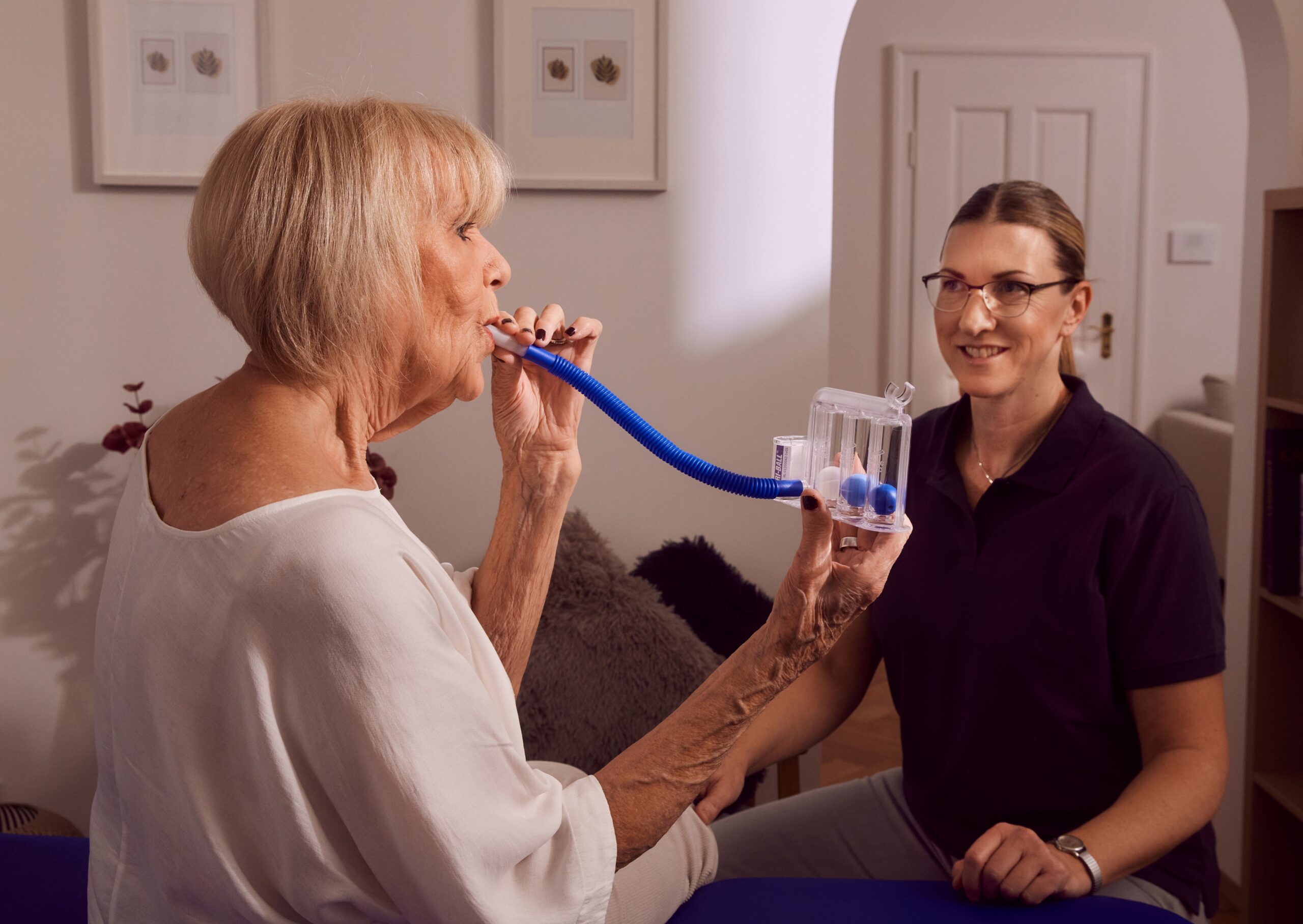 Ältere Frau übt Atemtherapie mit Gerät, betreut von Therapeutin.