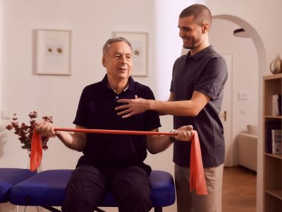 Älterer Mann trainiert mit Theraband, Physiotherapeut assistiert, in Praxis.