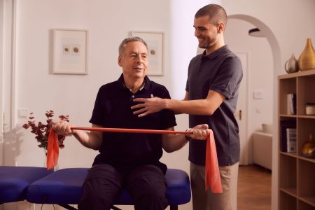 Älterer Mann trainiert mit Theraband, Physiotherapeut assistiert, in Praxis.