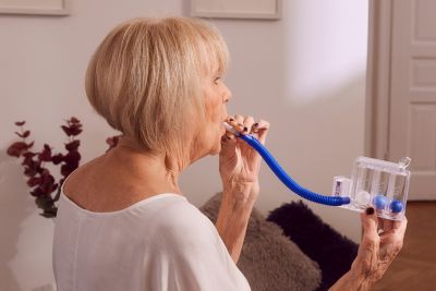 Patientin mit blonden Haaren im höheren Alter mit Lungenerkrankung trainiert mit Atemtrainingsgerät Triflo. Übungen in der Atemtherapie und Physiotherapie verbessern die Atmung. Patientin befinden sich zu Hause in ihrer Wohnung.