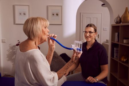 Ältere Frau übt Atemtherapie mit Gerät, betreut von Therapeutin.